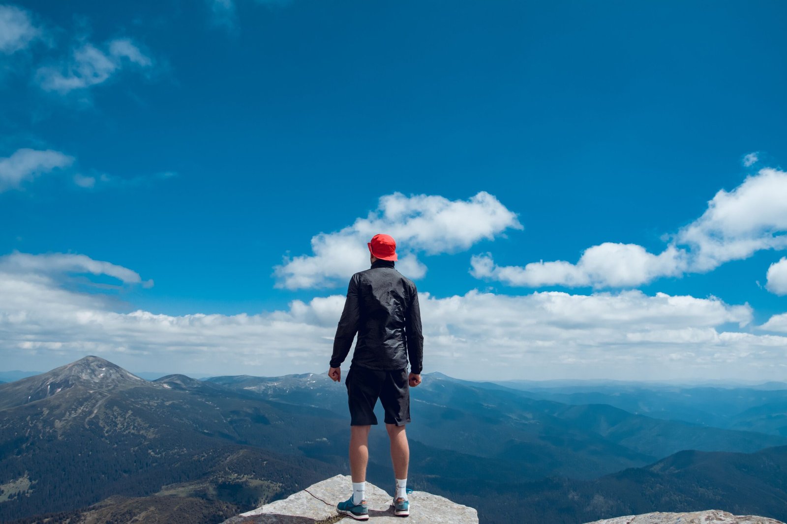 Man in red cap stands on cliff edge admiring Carpathian mountain range under clear blue sky.